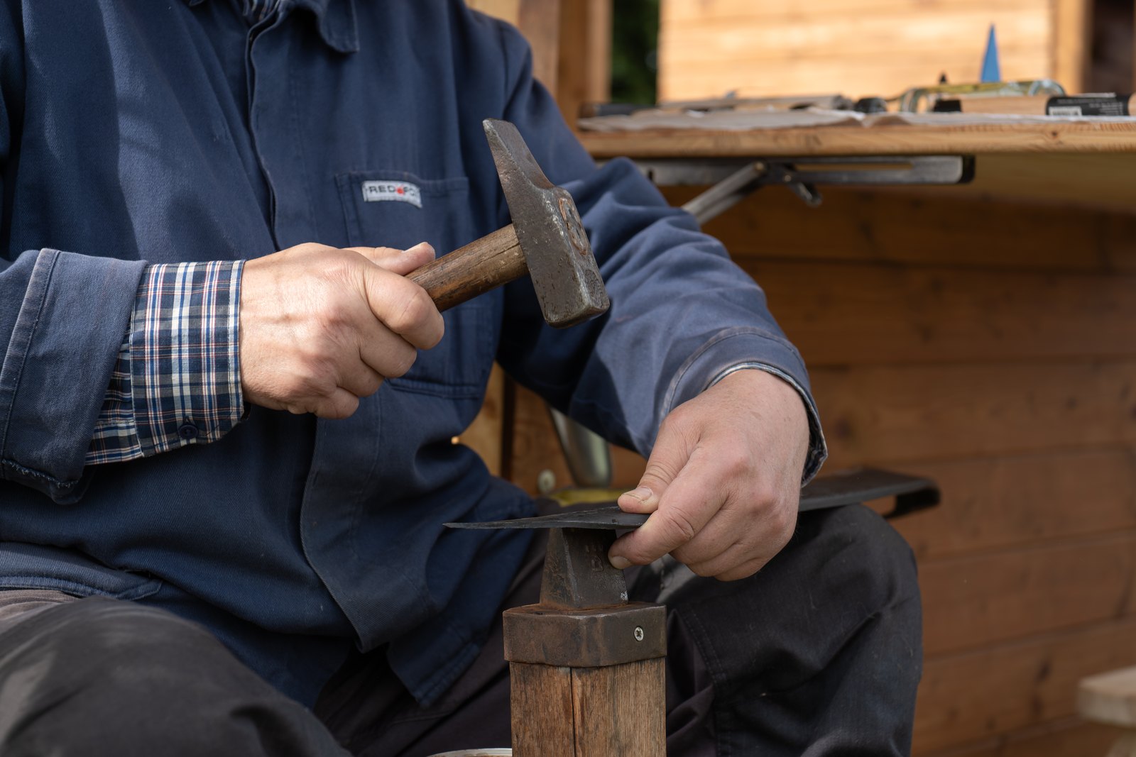 Hände eines Mannes in blauer Arbeitsjacke, der mit einem Hammer ein Sensenblatt auf einem Dengelamboss bearbeitet