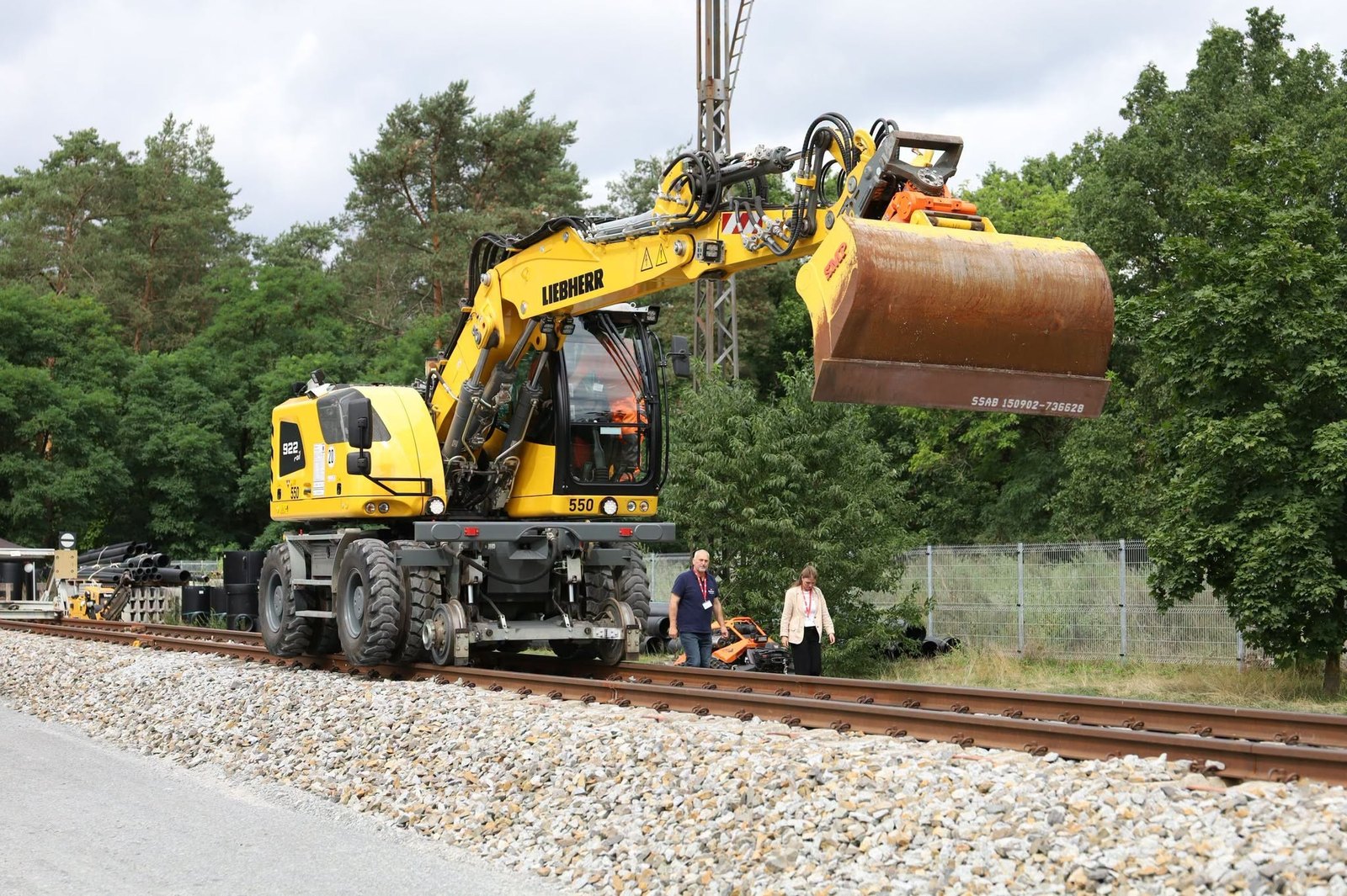 Gelber Zweiwegebagger mit angehobener Schaufel steht auf einem Gleis am Waldrand