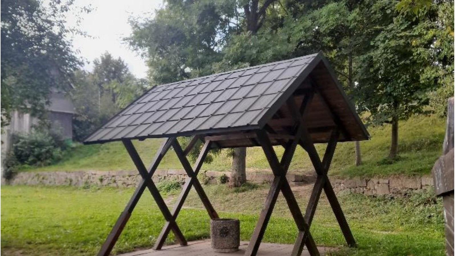 Ein hölzerner Unterstand mit Satteldach auf einer Wiese, darunter steht nur ein Papierkorb