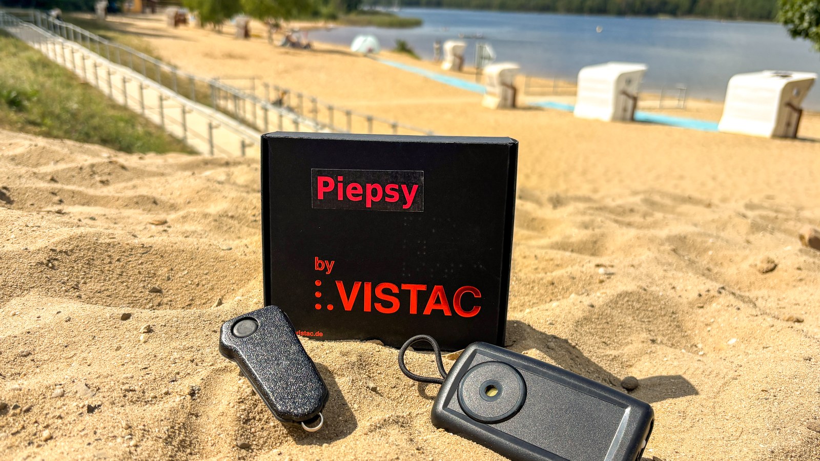 Zwei kleine schwarze Geräte und eine Verpackung stehen im Sand, dahinter Strandkörbe und ein See