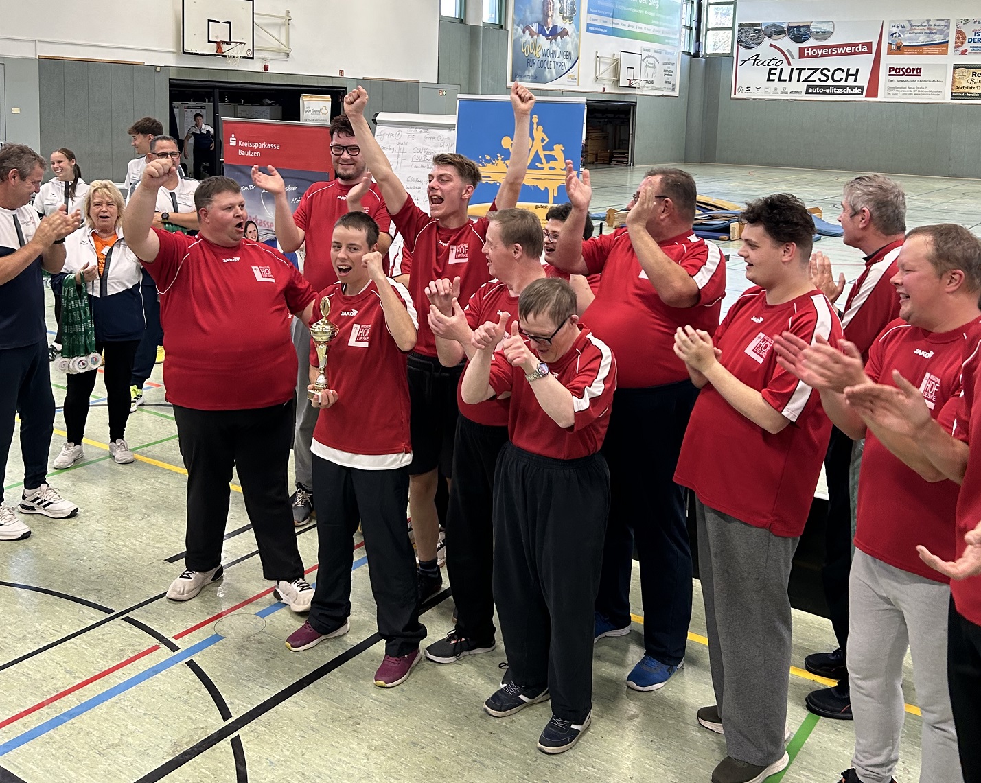 Eine Gruppe in roten Trikots jubelt in einer Sporthalle, einer hält einen Pokal