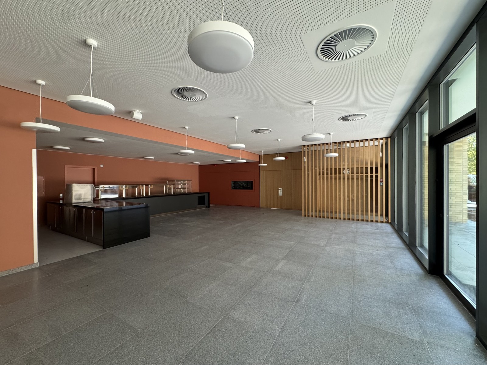 Heller Saal mit Fliesenboden, oranger Wandfläche, Holzlamellen und einer Ausgabetheke